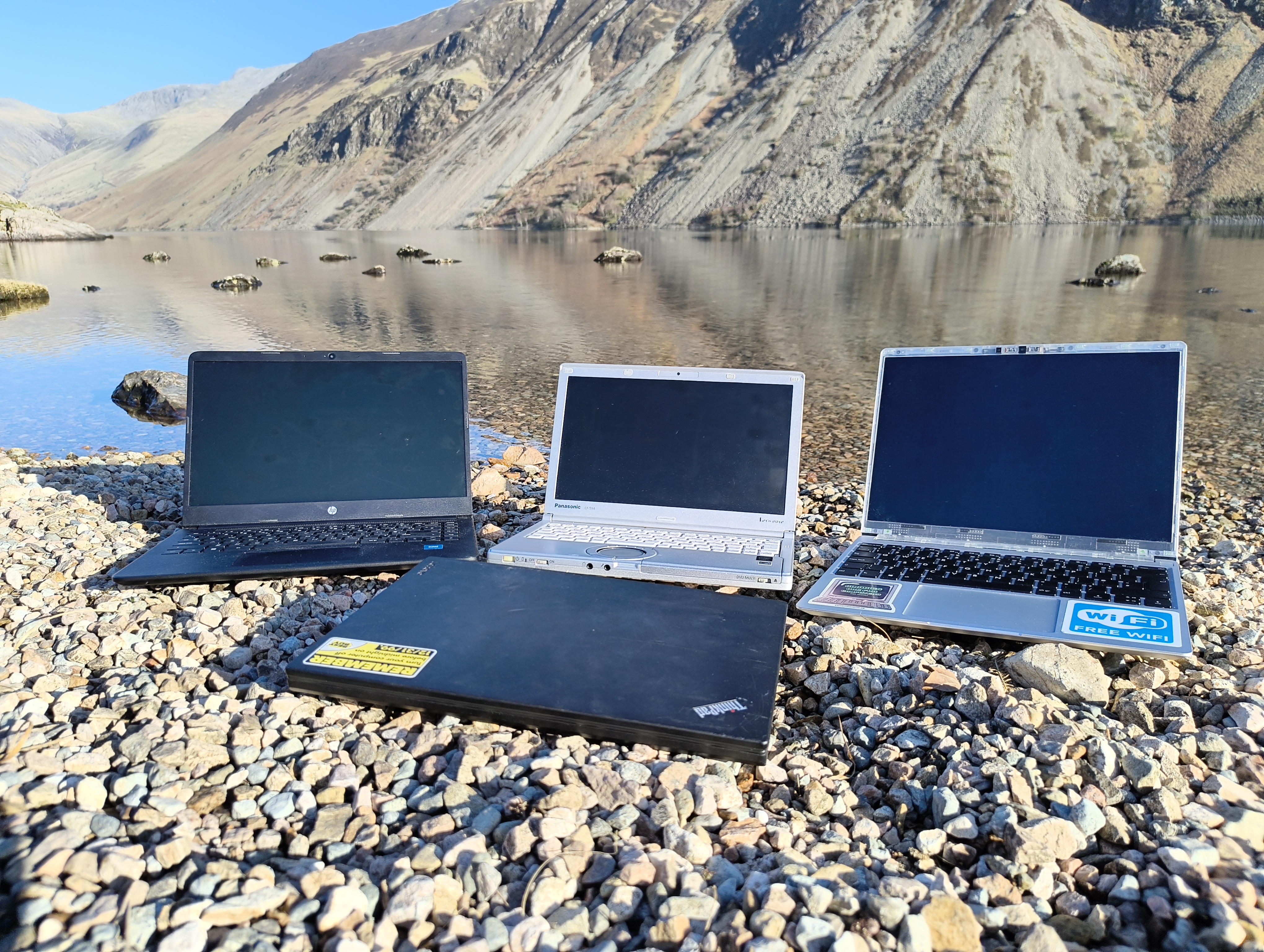 A Black Hp Laptop a Panasonic LetsNote a Framework Laptop and a Thinkpad x260 on a gravel beach with wastwater lake and the screes in the background